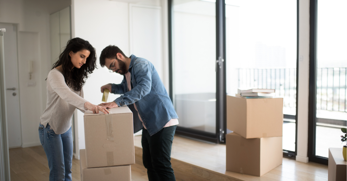 Couple packing boxes in a bright, modern apartment with large windows, preparing for a move