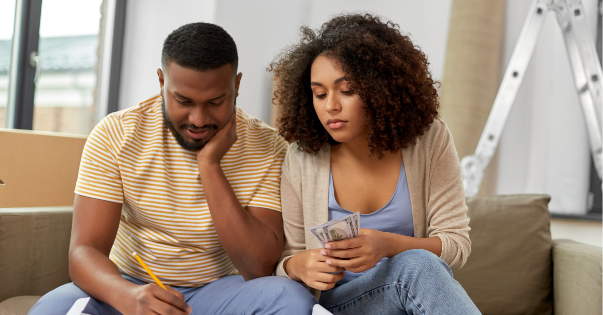 Couple sitting on a couch, calculating expenses with a notepad and holding cash, with moving boxes in the background.