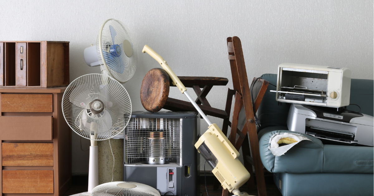 Collection of household items, including fans, a vacuum cleaner, a toaster, and furniture, piled together in a room