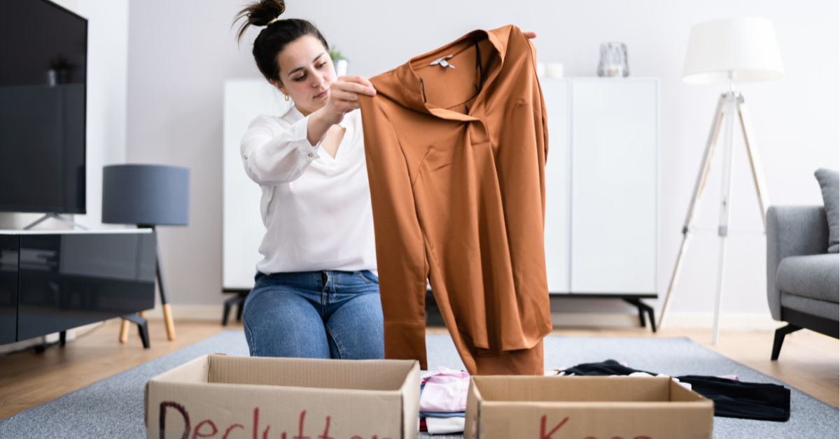 Woman holding up a brown blouse while sorting clothes into cardboard boxes labeled ‘Declutter’ and ‘Keep’ in a living room