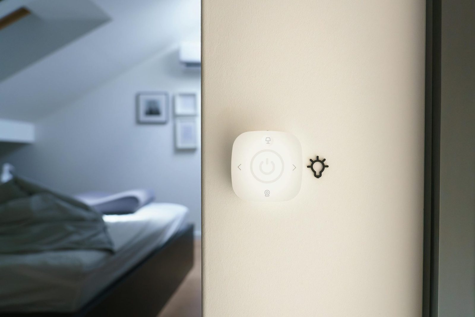 Smart home control panel mounted on a wall in a modern bedroom, with a blurred background of a neatly made bed and framed pictures on the wall