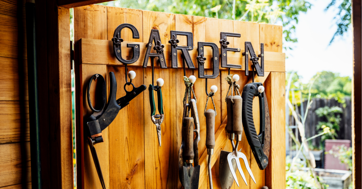 Garden tools hanging on hooks under a sign that reads ‘GARDEN’ on a wooden shed wall, with a garden view in the background