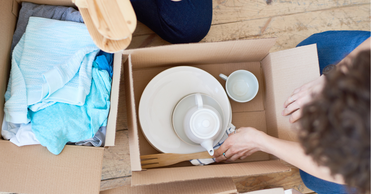 Person packing kitchen items, including plates and cups, into a cardboard box for moving, with another box containing folded clothes nearby