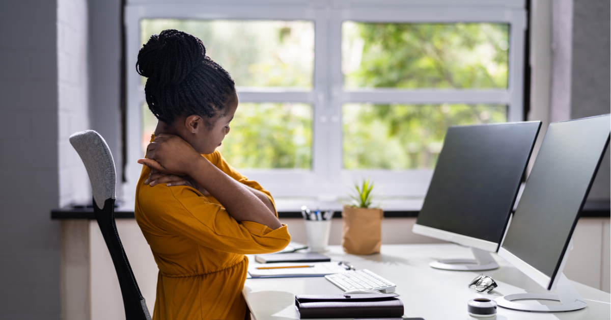 Woman stretching her neck and shoulders while sitting at an office desk with dual monitors, taking a break from work.