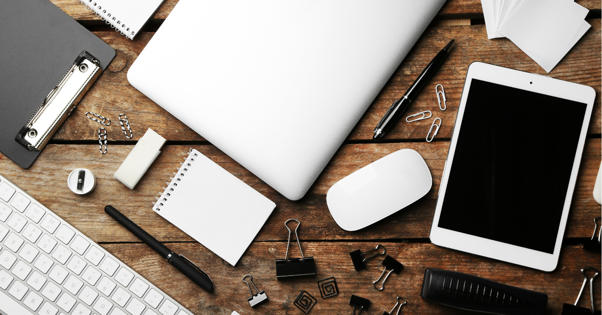 Flat lay of office supplies on a wooden desk, including a laptop, tablet, notepads, pens, paper clips, and a keyboard