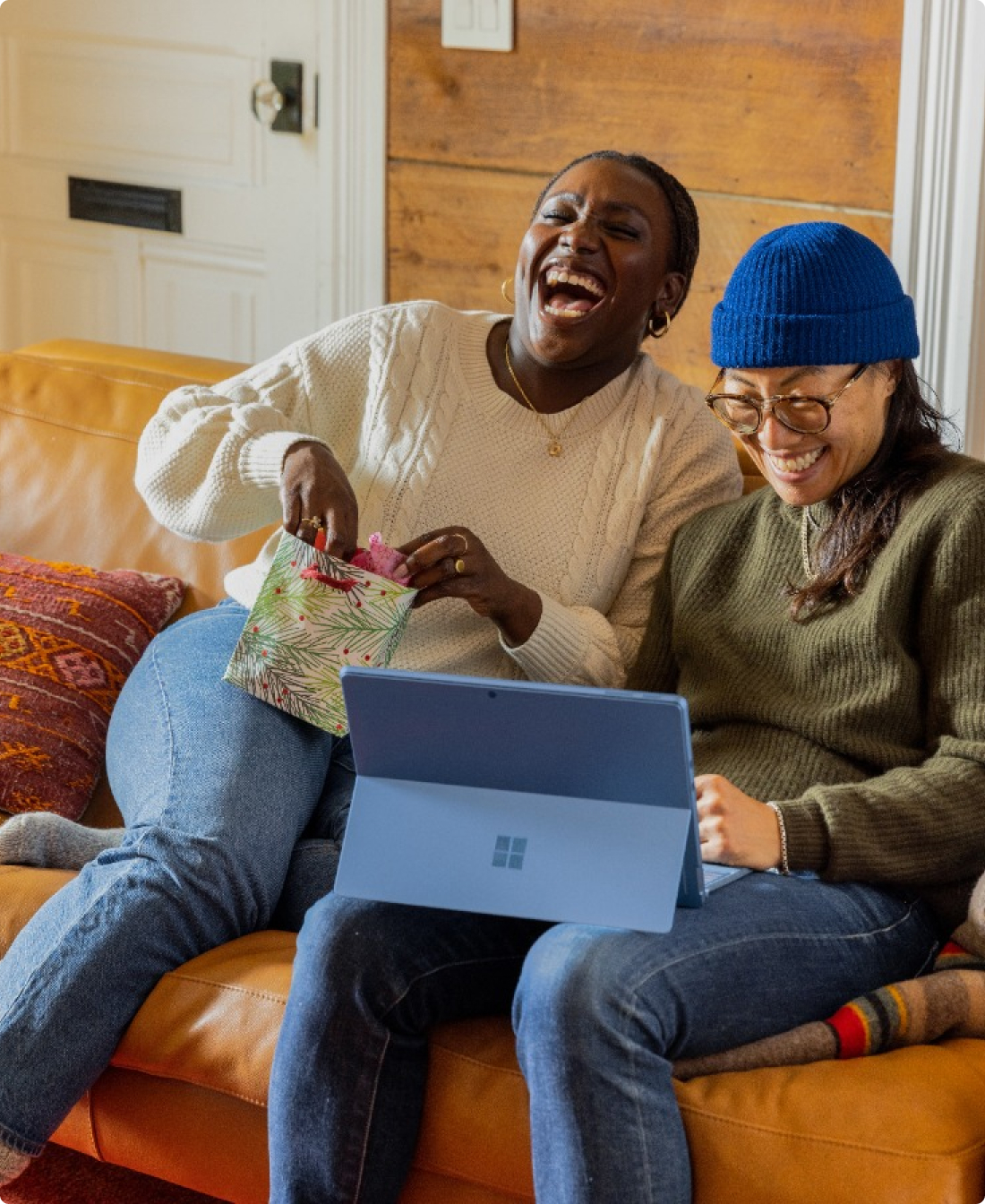 A couple of friends seating on a sofa laughing while they watch their tablet